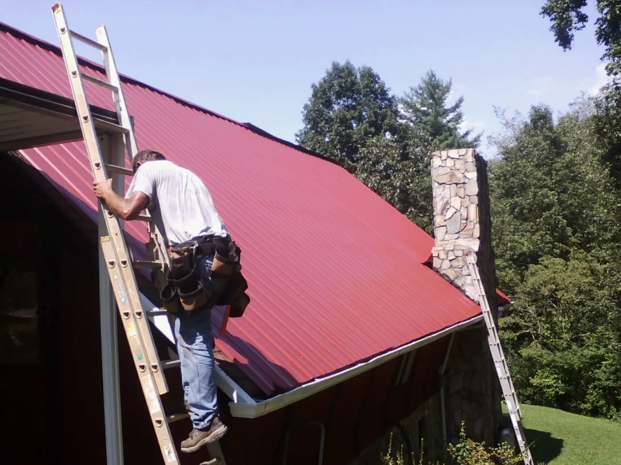 Roofer on a ladder beside a red metal roof and stone chimney — Soffit & Fascia Repair by Roofing Brothers in Bonner Springs